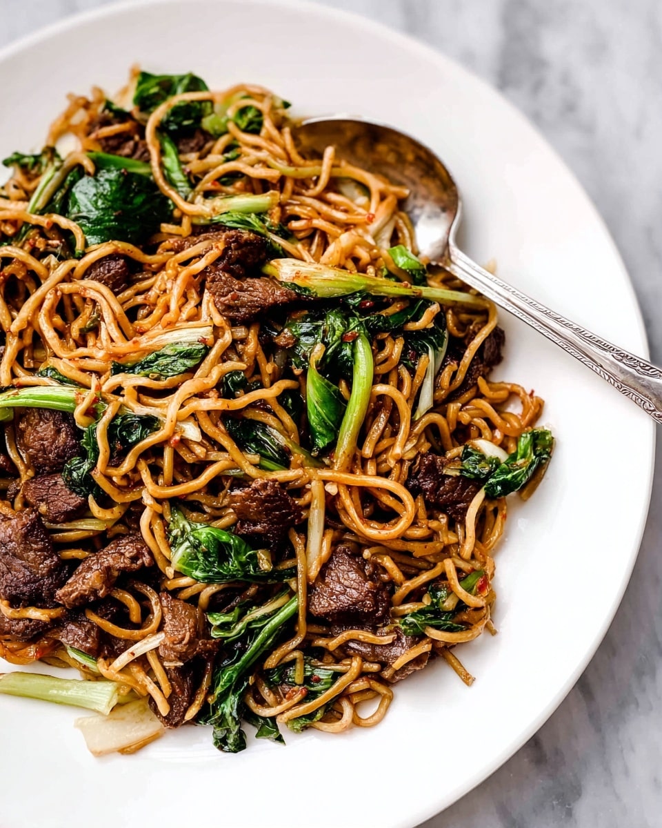 This dish shows a white plate filled with stir-fried noodles mixed with dark brown pieces of cooked meat and bright green leafy vegetables. The noodles are thin and a light golden brown color, twisted and spread out evenly among the meat and greens. The meat looks tender with a slightly crispy outside, while the green leaves have a fresh, slightly wilted texture, adding contrast in color. A silver spoon rests on the side, partly under the food in the plate. The plate is set on a white marbled surface. photo taken with an iphone --ar 4:5 --v 7