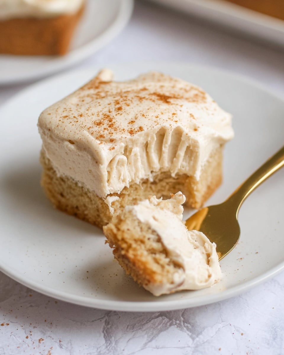 The image shows a close-up of a small dessert on a white plate with a white marbled texture background. The dessert has two layers: the bottom layer is a light brown, spongy cake, and the top layer is a thick, creamy, pale beige frosting that looks soft and fluffy, sprinkled lightly with cinnamon or another brown spice. A bite-sized piece of the dessert is held on a gold fork, showing the same two layers clearly. The focus is on the texture of the creamy top and the moist cake below. photo taken with an iphone --ar 4:5 --v 7