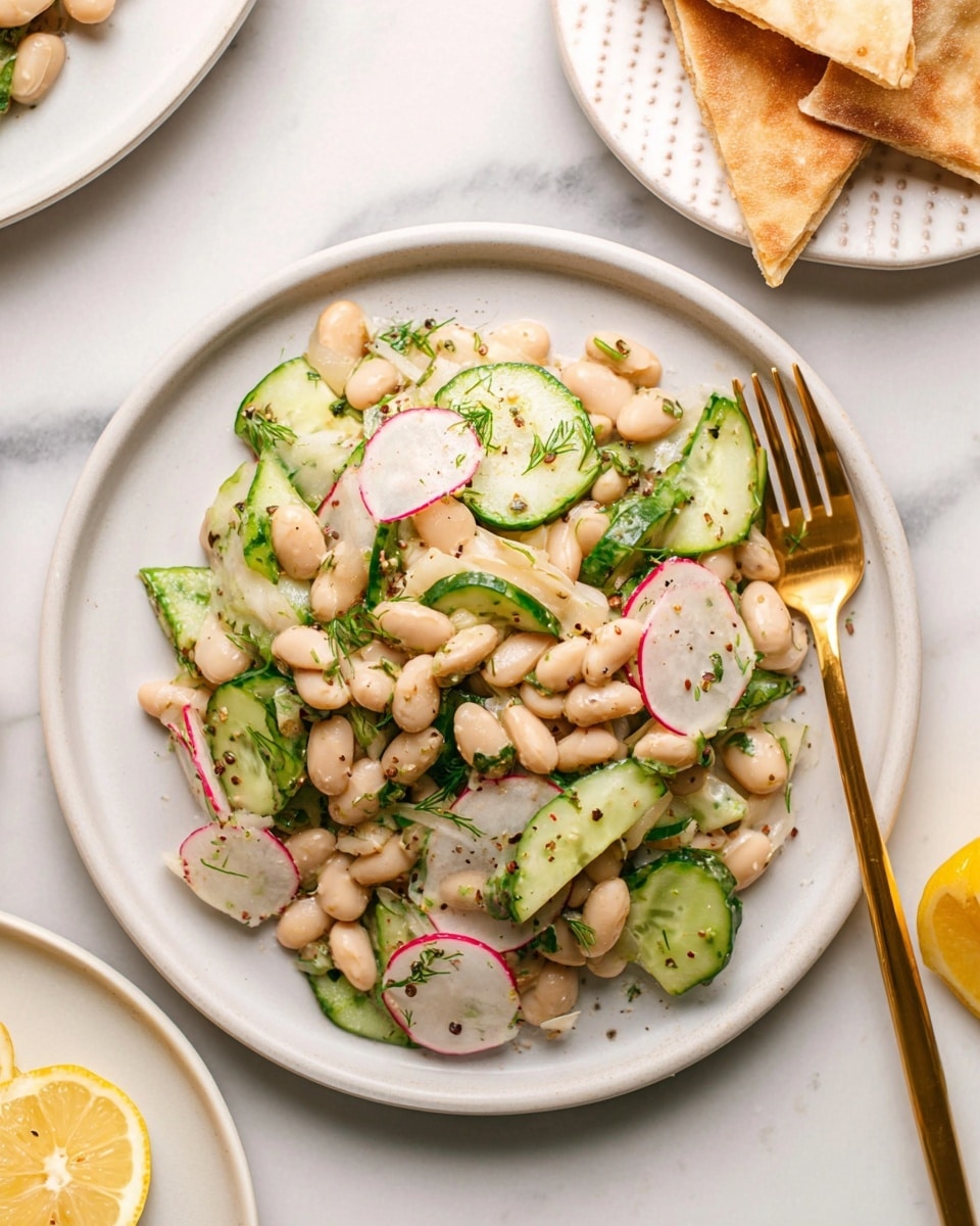 A white plate filled with a fresh salad made of light beige white beans, bright green cucumber slices, and thin white strips of radish with pink edges, all mixed with small green herb pieces and black pepper specks. The salad looks moist with a light dressing. A gold fork rests on the right side of the plate. In the background on the white marbled surface, there is a stacked pile of three white triangular flatbread pieces on a white plate and part of a lemon slice is visible on the right edge. photo taken with an iphone --ar 4:5 --v 7