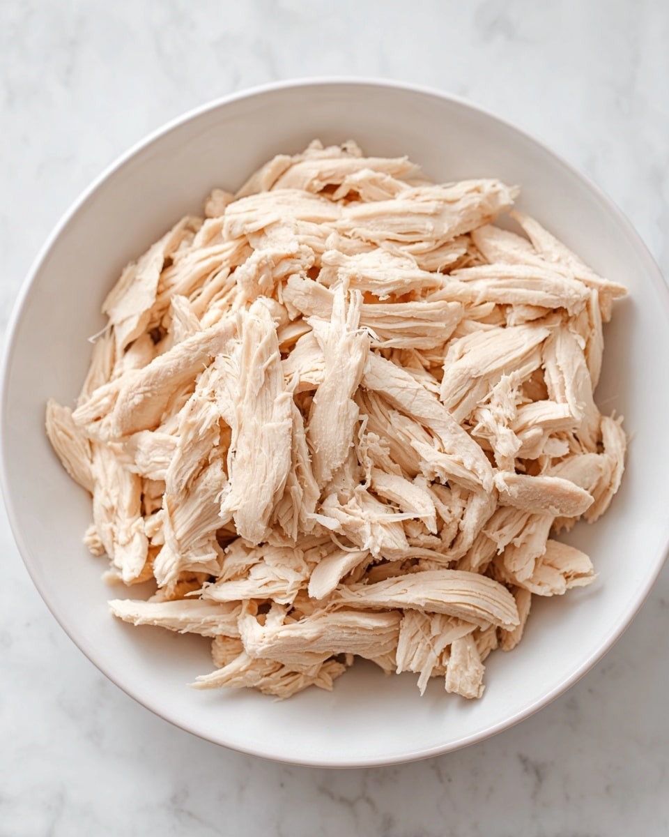 A white bowl filled with a single layer of shredded cooked chicken. The chicken pieces vary in size, with some slender and others thicker, all in a pale beige color showing the moist texture of the meat. The bowl sits on a white marbled surface, creating a clean and simple background. The focus is on the shredded chicken, neatly piled but not crowded, showing the fibrous, tender look of the meat. photo taken with an iphone --ar 4:5 --v 7