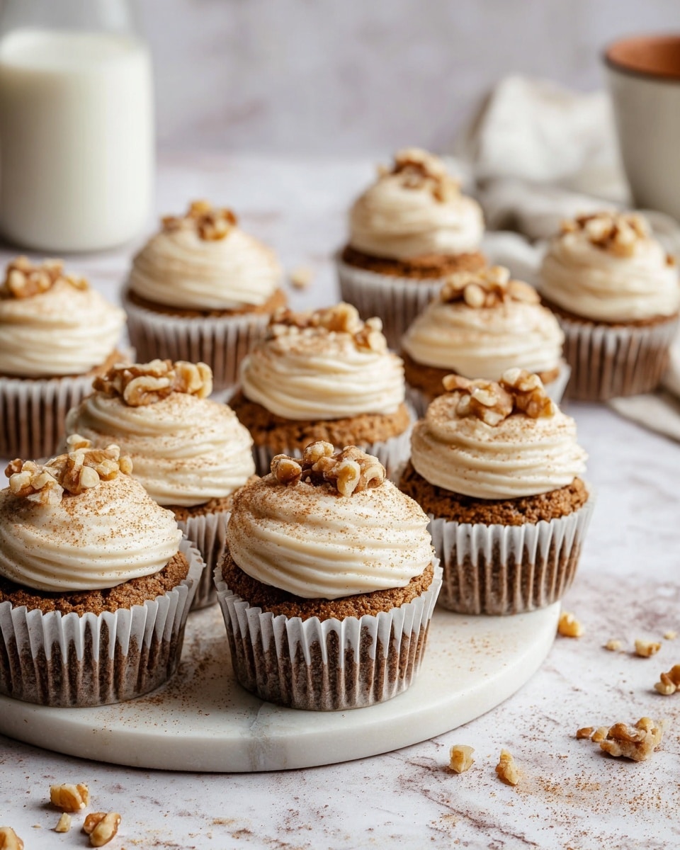 A group of twelve cupcakes arranged on a white plate with light gray marbling sits on a white marbled surface, each cupcake wrapped in white and brown striped paper. Each cupcake has two layers: a darker brown cake base with a soft texture and a thick layer of creamy white frosting swirled on top. The frosting is sprinkled lightly with brown cinnamon powder and topped with small, uneven pieces of light brown walnuts. Scattered walnut pieces and cinnamon powder are also spread on the white marbled surface around the plate. In the background, a tall glass of milk and a white and brown cup are partially visible. Photo taken with an iphone --ar 4:5 --v 7