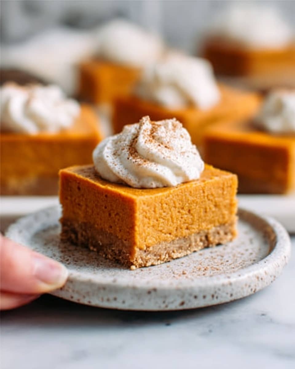 A close-up of a square piece of pumpkin pie with a thick, smooth orange filling and a crumbly light brown crust at the bottom. The pie has a small dollop of white whipped cream on top, slightly textured with soft peaks, and a sprinkle of light brown spice powder over the cream. It sits on a round white plate with grey marbled spots, resting on a white marbled surface. In the background, more pumpkin pie squares with whipped cream topper are blurred out. A woman's hand with a bite taken from the front side of the pie square is gently holding it. photo taken with an iphone --ar 4:5 --v 7