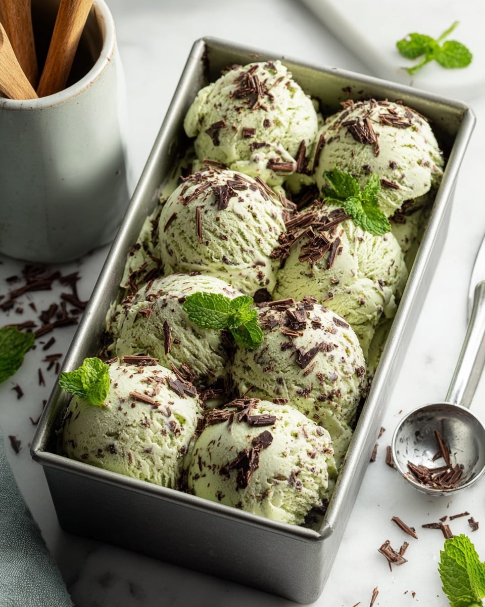 The image shows a rectangular metal container filled with eight scoops of pale green ice cream mixed with small dark chocolate pieces, giving a speckled texture. The scoops are layered close together, slightly uneven in shape, and topped with fresh bright green mint leaves and more dark chocolate shavings scattered on top. The container is placed on a white marbled surface, with a silver ice cream scoop beside it and some wooden spoons in a light gray jar in the background. Photo taken with an iphone --ar 4:5 --v 7