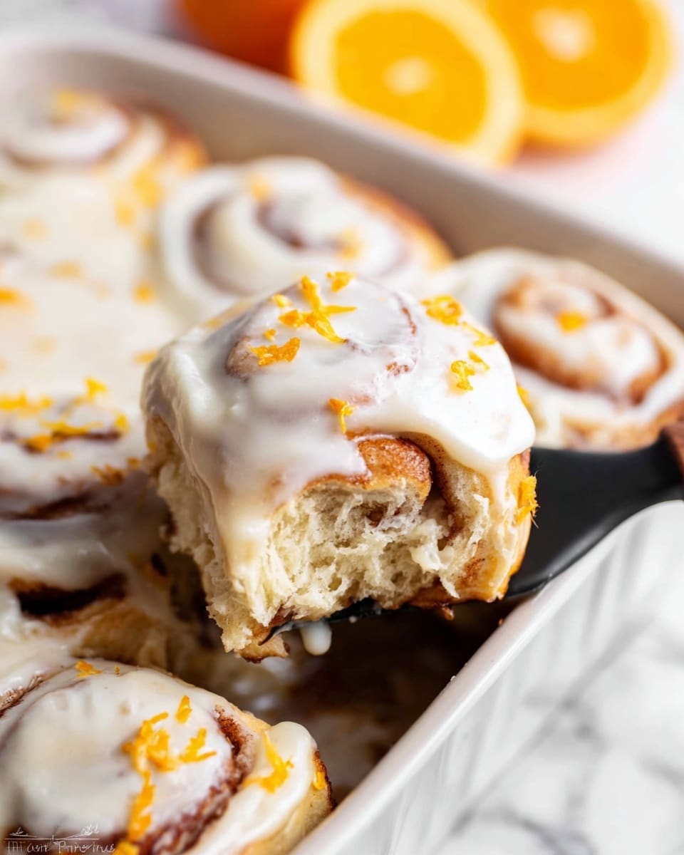 A close-up view of soft cinnamon rolls in a white rectangular baking dish filled with smooth, creamy white icing spread thickly over the top and sides. The cinnamon rolls show light golden-brown layers swirled with cinnamon beneath the icing, with small sprinkles of bright orange zest scattered unevenly on top, adding a pop of color. One cinnamon roll is being lifted by a black spatula, revealing the fluffy, light beige inside texture and swirled cinnamon layers. The background shows blurred halves of fresh oranges on a white marbled surface. photo taken with an iphone --ar 4:5 --v 7