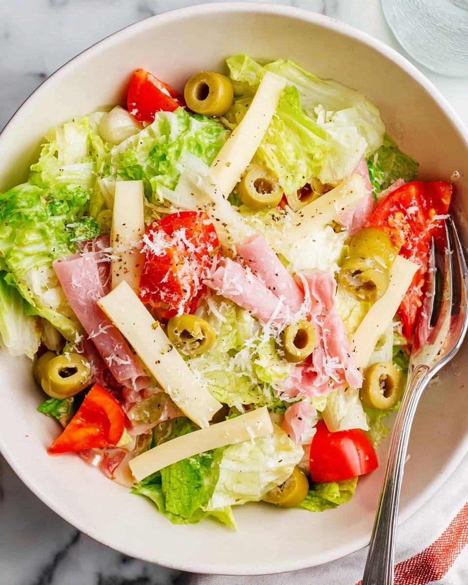 A white bowl filled with a fresh salad sits on a white marbled surface with a red and white cloth underneath. The salad has several layers: a base of light green lettuce leaves, topped with slices of pale yellow cheese sticks, thin pink slices of ham, wedges of red tomatoes, and green olives with pimento. Shredded white cheese is sprinkled over the top, and a silver fork is placed inside the bowl, partially immersed in the salad. In the blurred background, a wooden bowl holds more chopped salad ingredients. Photo taken with an iphone --ar 4:5 --v 7