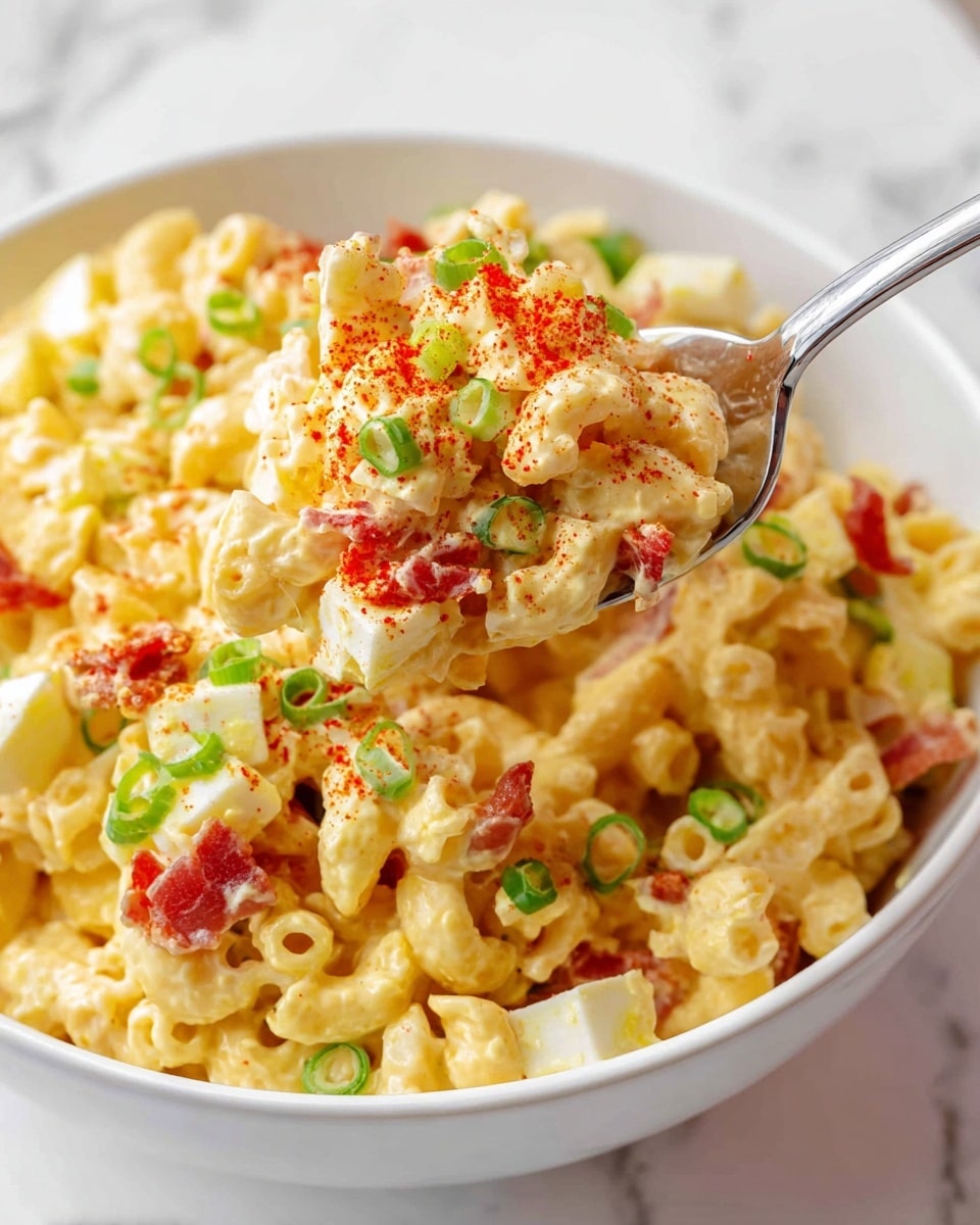 A white bowl filled with a creamy macaroni salad showing small elbow macaroni layered with a smooth, yellowish cheese sauce mixed with diced pale yellow eggs and bits of red bacon. The salad is sprinkled with finely chopped green onions and a light dusting of red paprika. A shiny silver spoon lifts a portion of the salad, highlighting the different textures and colors against a white marbled surface. photo taken with an iphone --ar 4:5 --v 7