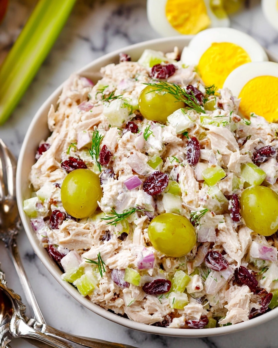 A close-up view of a creamy chicken salad served in a white bowl, filled with layers of shredded white chicken mixed with small pieces of light green celery, diced red onion, and bits of dark red dried cranberries. Scattered green grapes, sliced in half, add a fresh yellow-green color, while small pieces of chopped green onions and sprigs of dill lightly top the salad. Two halved boiled eggs with bright yellow yolks sit near the center on top. The bowl is placed on a white marbled surface with a celery stick and silver serving utensils nearby. Photo taken with an iphone --ar 4:5 --v 7