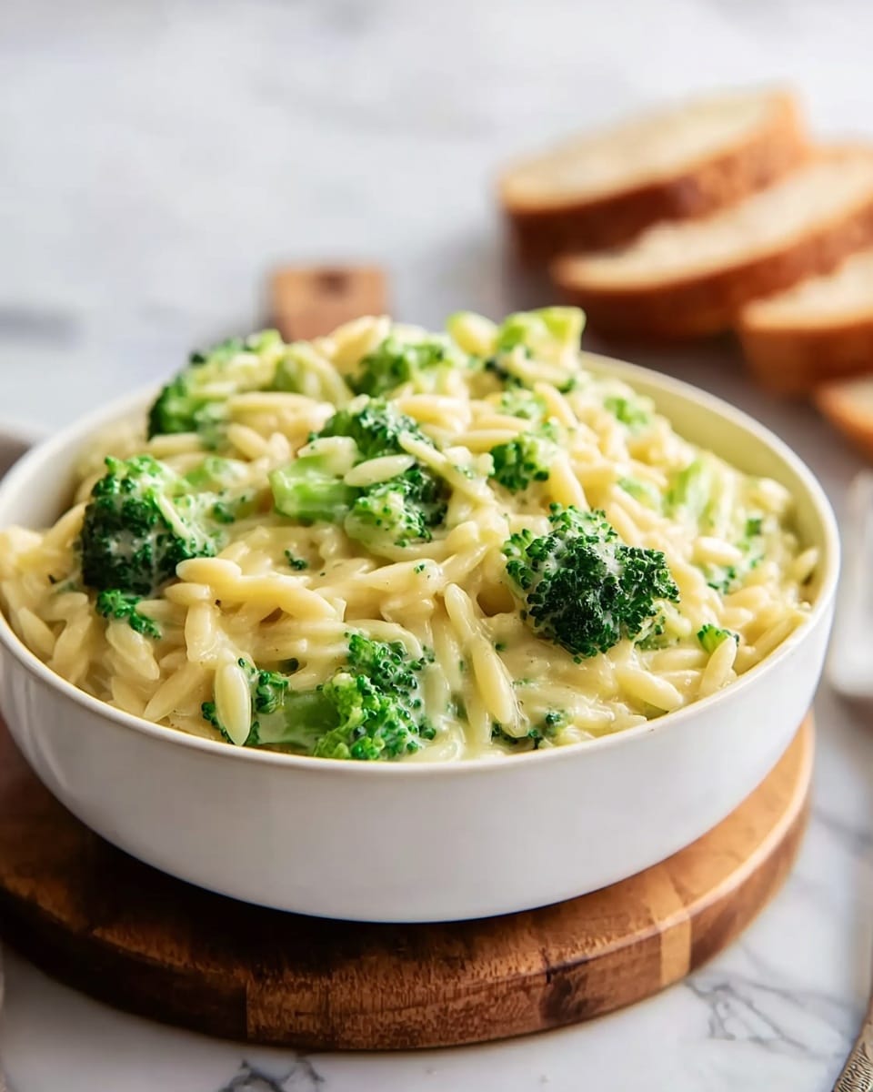 A white bowl filled with a creamy pasta dish sits on a round wooden board placed on a white marbled surface. The dish has two main layers: at the bottom, soft orzo pasta in a light yellow color is coated with a creamy sauce, and mixed throughout are bright green broccoli florets. The texture looks smooth and creamy, with the broccoli adding some contrasting roughness and green color. In the blurred background, there are slices of bread. The photo taken with an iphone --ar 4:5 --v 7