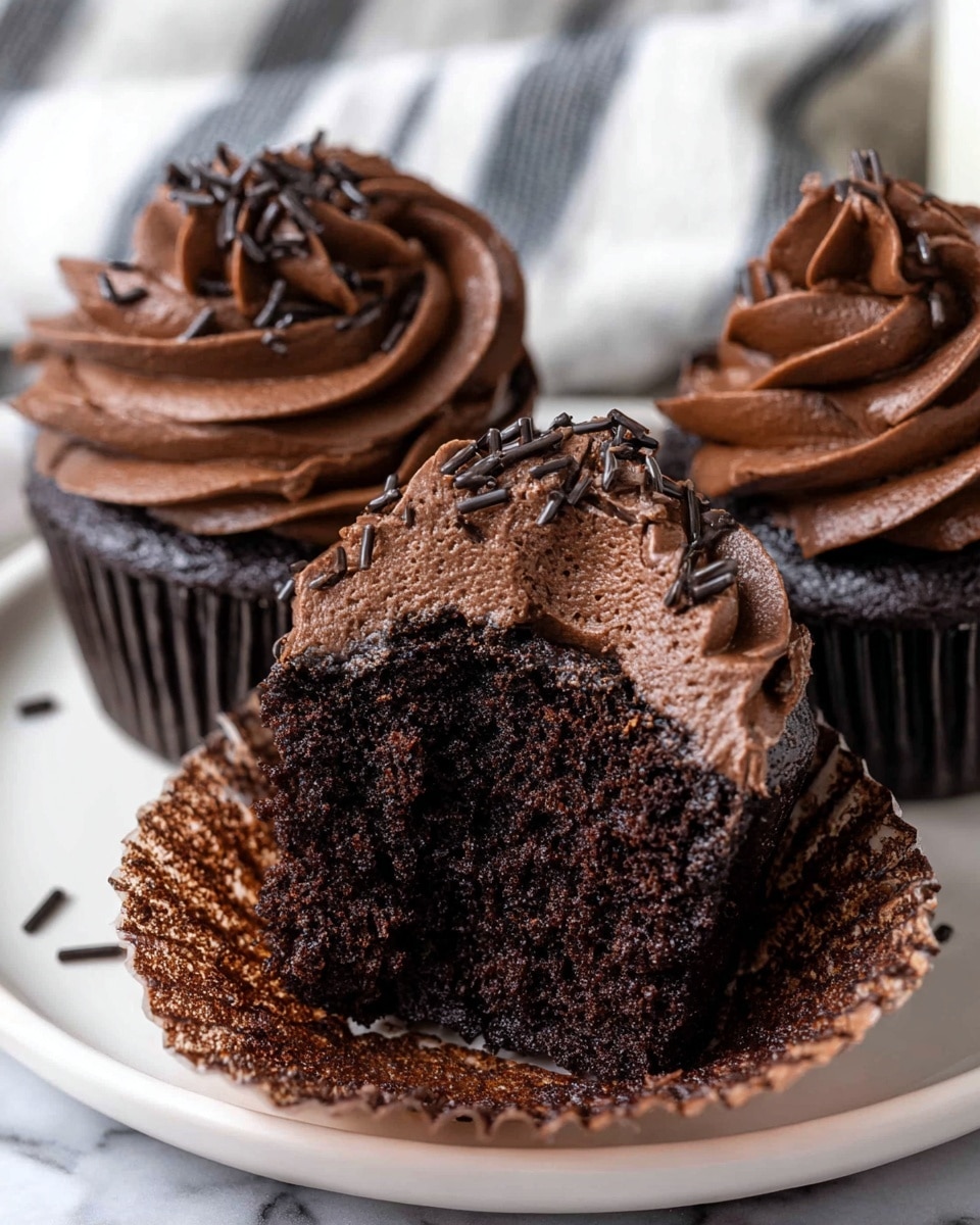 A close-up of three dark chocolate cupcakes on a white plate with a white marbled surface underneath, each topped with a large swirl of thick, rich dark chocolate frosting that is shiny and smooth with a slightly wavy texture. The frosting is decorated with small, thin, black sprinkles scattered on top. One cupcake is cut in half showing a moist, dense, and deep brown chocolate cake inside with a slightly crumbly texture. The background has a soft focus with a white and black striped cloth visible. Photo taken with an iphone --ar 4:5 --v 7