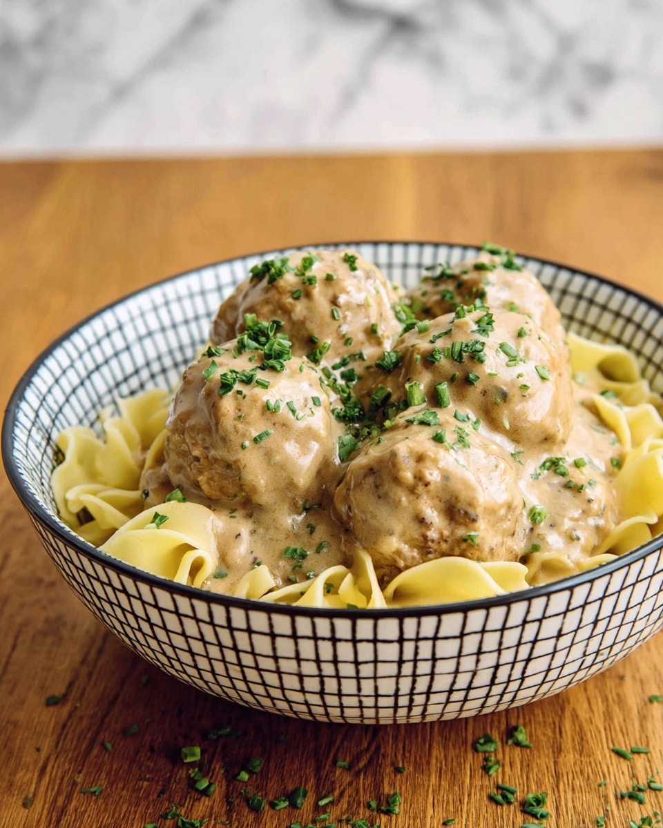 The image shows a bowl with a white base and a black square pattern, filled with three layers: at the bottom, there is a layer of yellow egg noodles with a slightly ruffled texture; above the noodles, there are large, round meatballs with a rough, textured surface coated with a thick, creamy, light brown gravy; the top layer includes small chopped green herbs sprinkled over the meatballs and gravy. The bowl sits on a wooden table with some scattered green herbs around it, and the background is a white marbled texture. Photo taken with an iphone --ar 4:5 --v 7