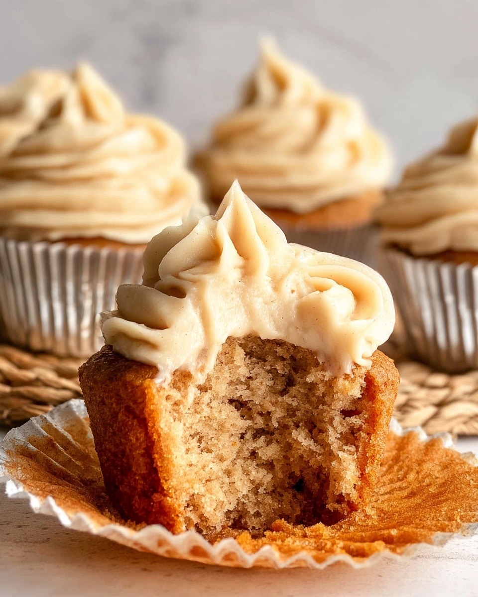 A close-up of a single cupcake with a bite taken out of it, showing its soft, crumbly light brown cake inside as the bottom layer. The top layer is creamy beige frosting with a swirled, smooth texture, generously piped on. Behind it, three whole cupcakes with the same frosting and cake color sit on a light brown woven mat, all wrapped in silver foil liners. The background is a white marbled texture. photo taken with an iphone --ar 4:5 --v 7