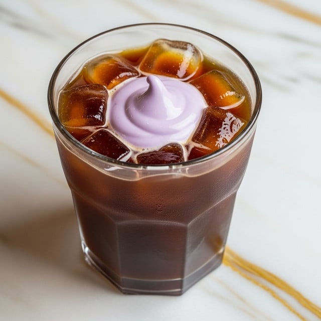A clear glass filled with dark brown iced coffee with several large ice cubes floating inside. On top, there is a smooth, thick swirl of light purple cream that covers part of the ice and coffee, creating a soft creamy texture with a gentle peak in the center. The glass sits on a white marbled surface with gold veins. photo taken with an iphone --ar 4:5 --v 7