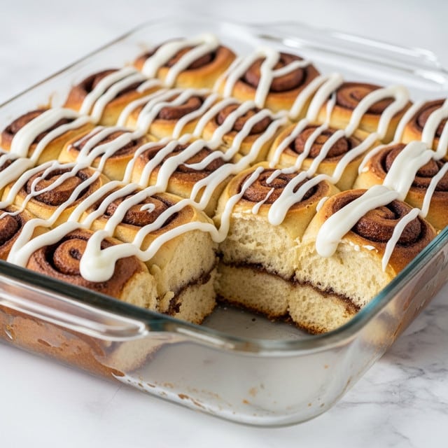 A glass baking dish filled with tightly packed, golden-brown cinnamon roll pieces, each one showing a slightly toasted, crispy surface with darker caramelized edges. The rolls are drizzled unevenly with thick white icing, some pooling around the base, creating a glossy, sticky texture on top. The dish sits on a white marbled surface that highlights the warm tones of the rolls, emphasizing their soft interior and well-baked crust. Photo taken with an iphone --ar 4:5 --v 7