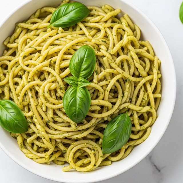 A close-up of a bowl full of spaghetti pasta twisted into small piles, coated evenly with a green pesto sauce that has a slightly oily and textured look with tiny bits of herbs and nuts visible. Two fresh green basil leaves are placed on top and mixed within the pasta, adding freshness and bright color contrast. The white bowl sits on a white marbled surface, giving a clean and simple background. The photo taken with an iphone --ar 4:5 --v 7
