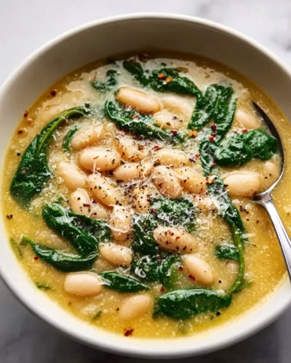 A white bowl filled with a thick soup, showing three main layers: the bottom layer is a light orange, creamy broth; on top of that are large, soft white beans spread evenly; the upper layer consists of dark green leaves, likely spinach, scattered around the beans. Small bits of grated cheese are sprinkled over the surface, adding texture, and a silver spoon rests inside the bowl on the right side. The bowl sits on a white marbled surface. Photo taken with an iphone --ar 4:5 --v 7