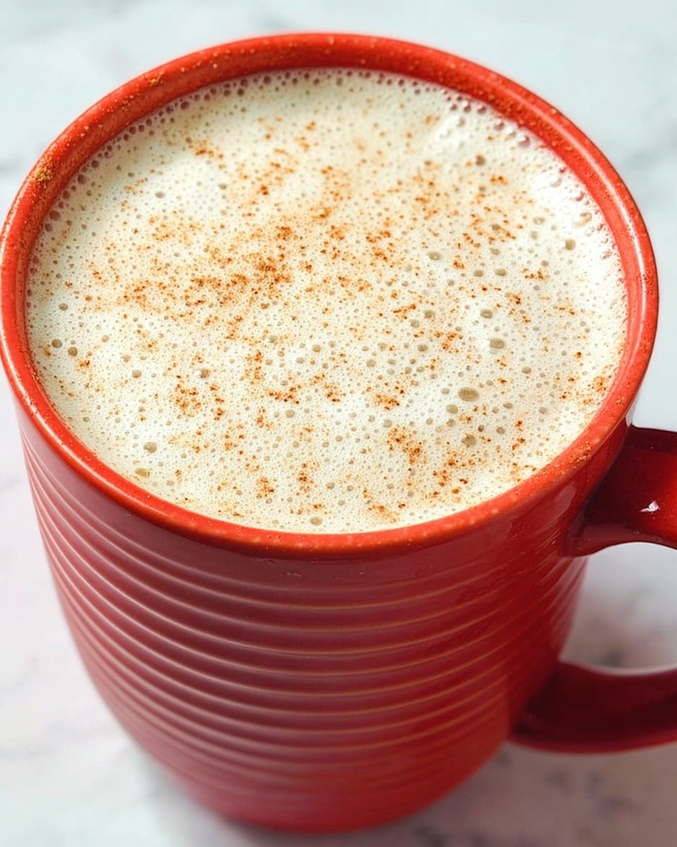 A close-up view of a bright orange cup filled with a creamy frothy drink topped with a light sprinkle of brown spice, likely cinnamon or nutmeg. The drink has a smooth foam layer with small bubbles covering the surface evenly. The cup is set against a white marbled texture background, and the handle is visible on the right side. photo taken with an iphone --ar 4:5 --v 7