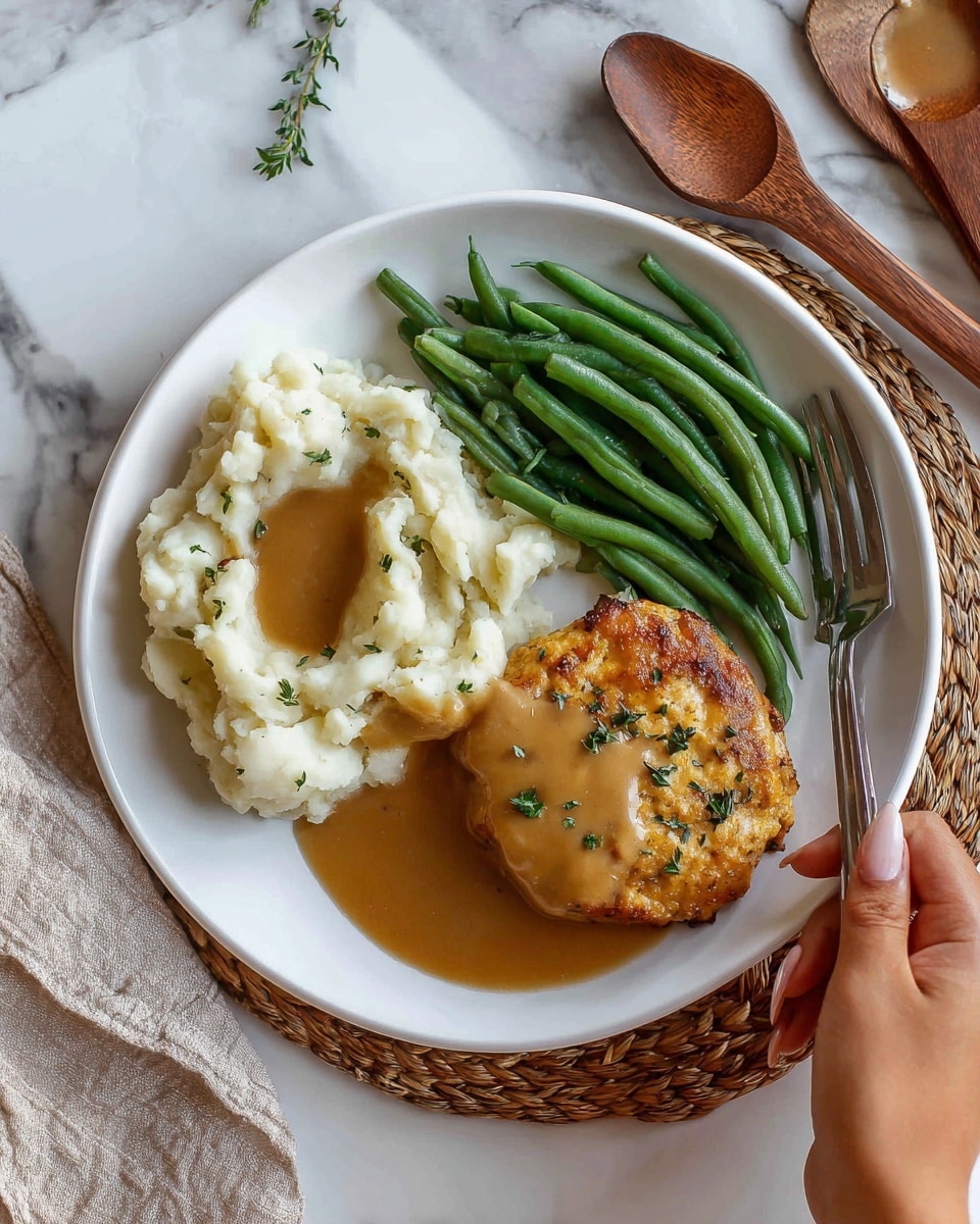 A white plate on a white marbled surface holds a meal with three main parts: on the left, smooth, creamy mashed potatoes topped with a light brown gravy; at the top, bright green cooked green beans; on the right, a grilled, golden-brown patty sprinkled with small green herbs, all sitting in a pool of thick, tan-colored sauce. There is a silver fork and knife to the right of the plate. Photo taken with an iphone --ar 4:5 --v 7