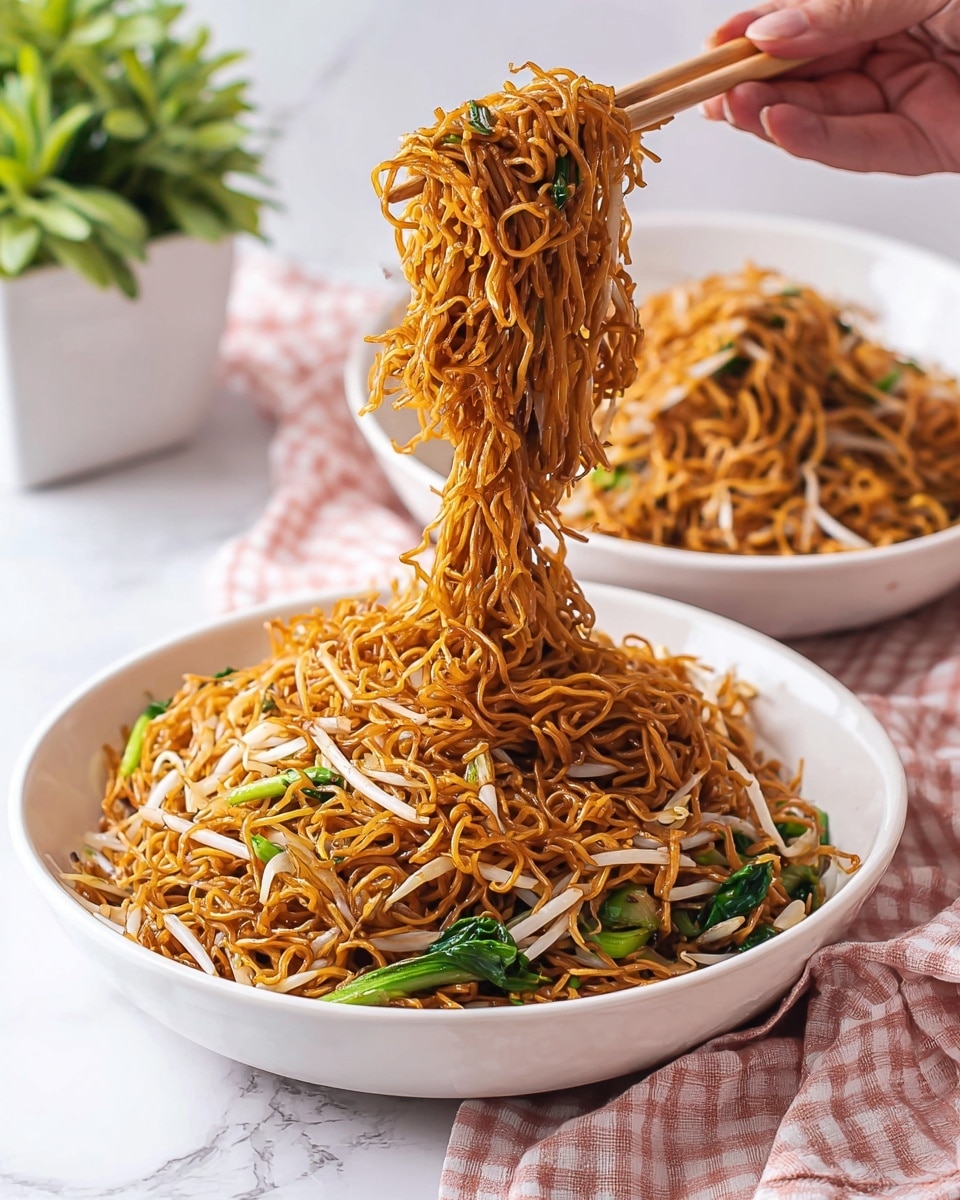 A white shallow bowl filled with a heap of dark golden-brown fried noodles mixed with light beige bean sprouts and green leafy vegetables. A woman's hand is holding light pink chopsticks lifting a clump of the thin, shiny noodles, stretching them above the bowl. In the background, there is another white bowl with more noodles, and a potted green plant with broad leaves. All items are placed on a white marbled surface with a pink and white checkered cloth at the bottom edge of the image. Photo taken with an iphone --ar 4:5 --v 7