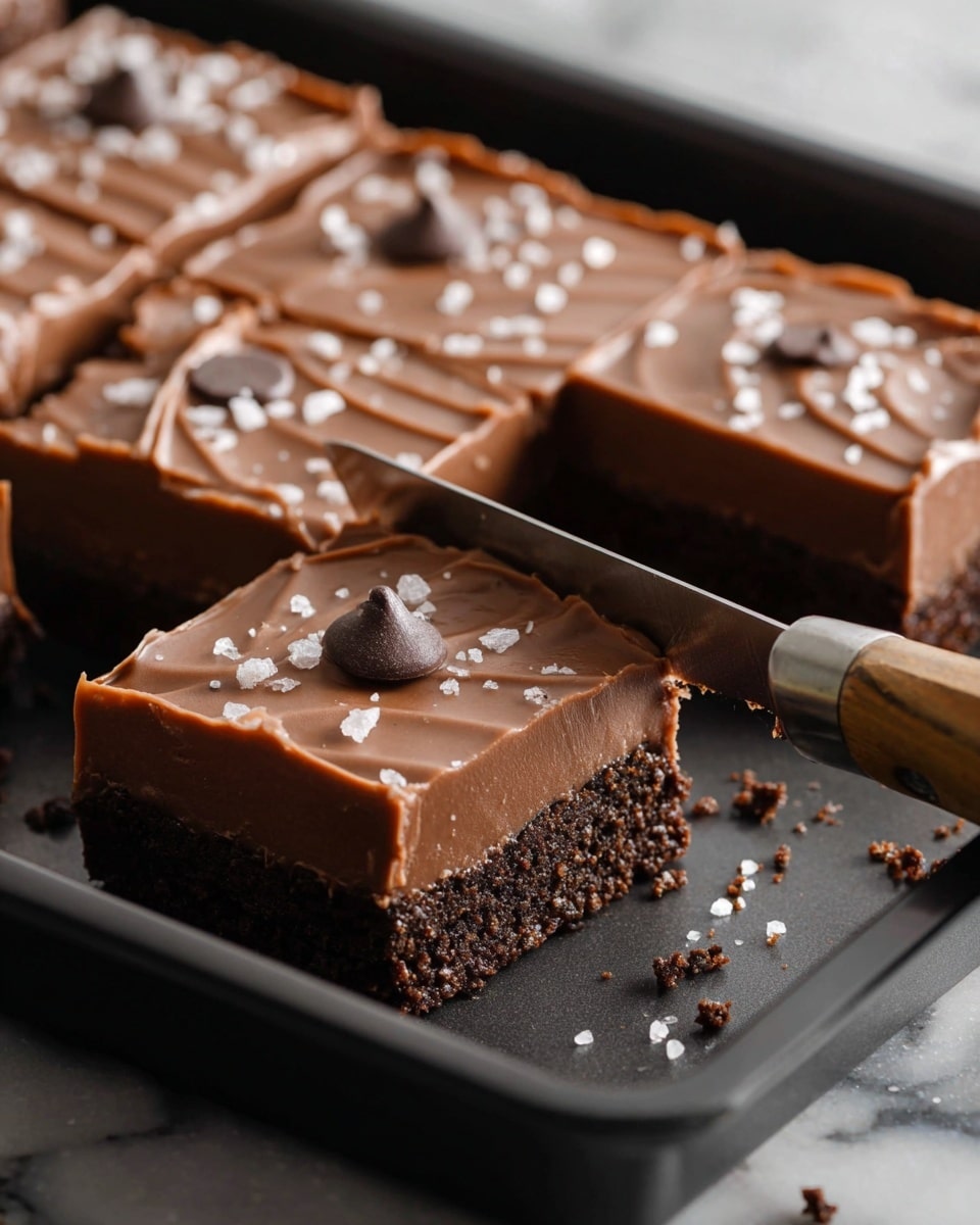A close-up view of chocolate squares on a dark baking tray, each piece topped with smooth milk chocolate icing, lightly rippled on top, and sprinkled with coarse white sea salt and a single dark chocolate chip in the center. The bars have two visible layers: a thick, glossy milk chocolate top layer and a dense, darker chocolate base layer, with a crumbly crust that looks slightly textured and rough around the edges. A knife with a wooden handle is cutting through one of the squares, showing the clean separation between layers. The scene is set on a white marbled surface with some chocolate crumbs and salt flakes scattered around. photo taken with an iphone --ar 4:5 --v 7