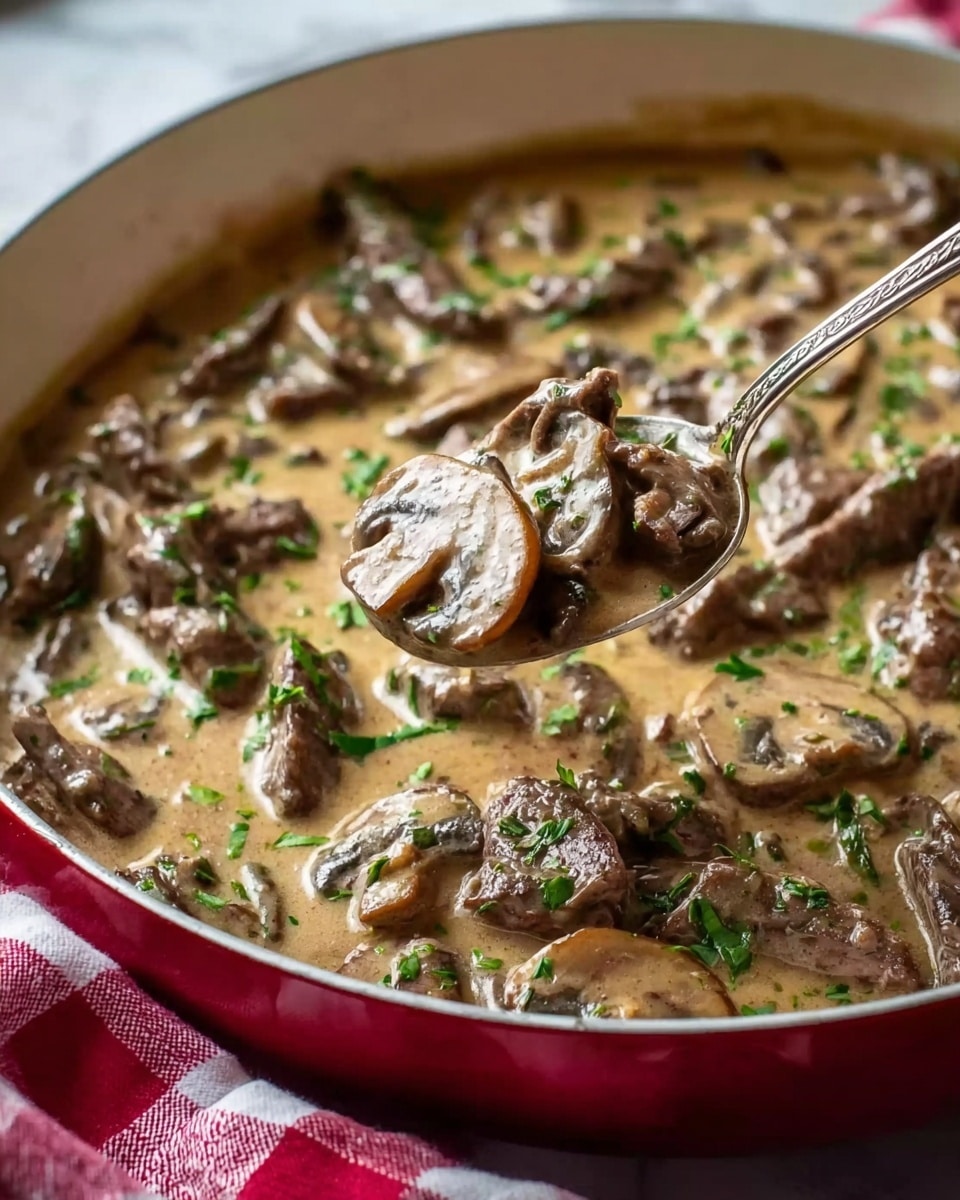 The image shows a close-up of a creamy mushroom and beef stew in a white pan with a red outside edge. The dish has several layers: thin slices of brown mushrooms and tender pieces of cooked beef in the bottom layer, covered by a light brown creamy sauce with small green herb pieces sprinkled on top. A silver spoon is lifting a spoonful of the creamy mixture that includes both mushroom slices and beef, showing the rich texture of the dish. The pan sits on a white marbled surface with a red and white checkered cloth partly visible on the side. photo taken with an iphone --ar 4:5 --v 7
