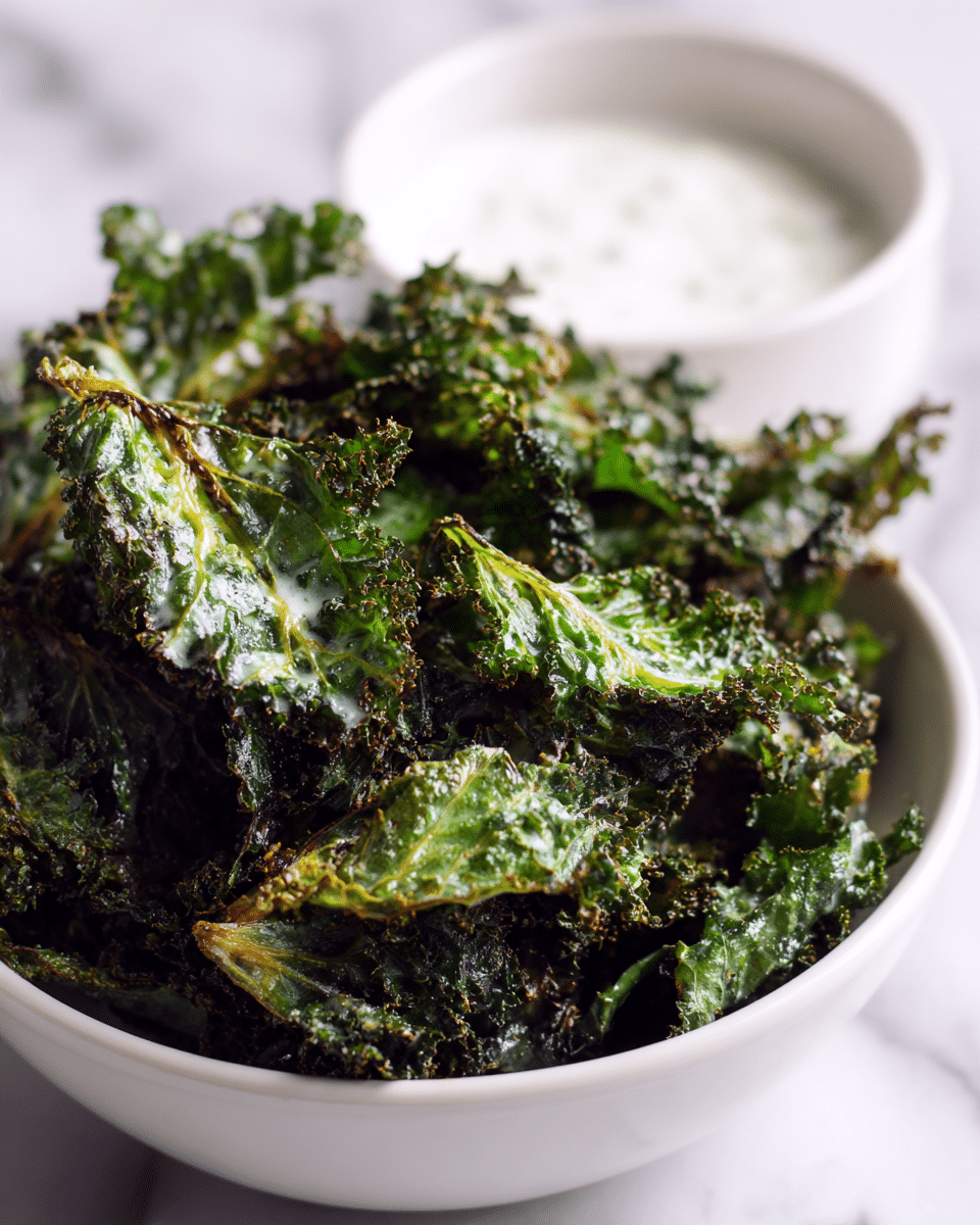 The image shows a white bowl full of crispy kale chips, which are dark green with a rough, curly texture. The kale chips look crunchy and slightly shiny, as if they are lightly coated with oil. A small white bowl with a creamy white dip is placed in the background, slightly out of focus. The setting is on a white marbled texture surface that adds a clean, bright look to the image. photo taken with an iphone --ar 4:5 --v 7