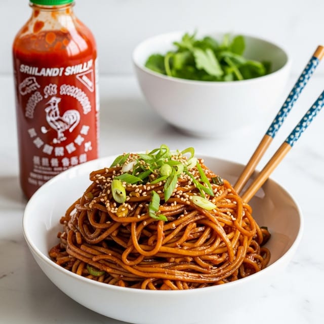 A white bowl filled with glossy, dark reddish-brown noodles layered thickly and mixed with small bits of vegetables and sauce, topped with bright green sliced scallions and light tan sesame seeds; two chopsticks with blue patterned tips rest inside the bowl on the right side. In the background, a bottle of red-orange sriracha chili sauce with a yellow dragon design stands tall on a white marbled surface, alongside a white bowl containing fresh green herbs. The overall image has bright light and clean presentation. photo taken with an iphone --ar 4:5 --v 7