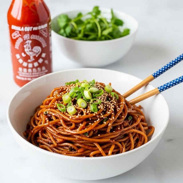 A white bowl filled with thick, dark reddish-brown noodles coated in sauce, garnished with green sliced scallions and sprinkled with white sesame seeds on top. Two yellow chopsticks with a dark blue and white pattern rest inside the bowl, leaning on the side. Behind the bowl, a bottle of sriracha chili sauce with a bright orange and yellow label is placed on a white marbled surface, along with a white bowl containing fresh green herbs. The overall look is warm and appetizing with a neat, clean setup. photo taken with an iphone --ar 4:5 --v 7
