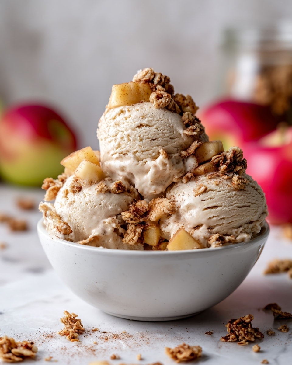The image shows a white bowl filled with three scoops of light beige ice cream, each scoop smooth and creamy. On top and around the ice cream are small chunks of cooked apples with a warm golden-brown color, lightly coated with cinnamon, adding texture. Sprinkled over the ice cream and apples are clusters of granola, dark golden and crunchy, which contrast with the soft ice cream. The white bowl rests on a white marbled surface. In the blurry background, there are colorful apples and a jar with a blurred label, providing a warm and fresh feel to the scene. Photo taken with an iphone --ar 4:5 --v 7