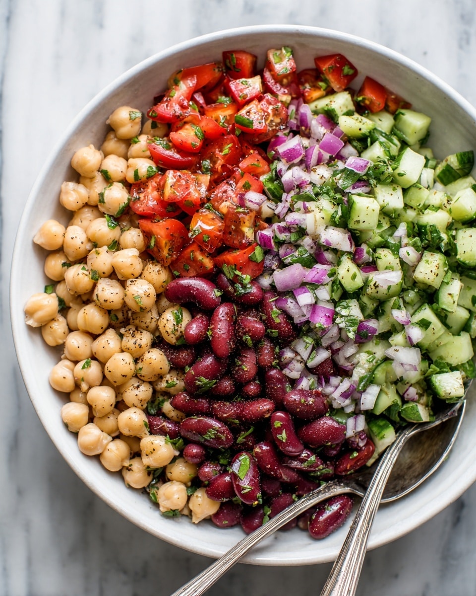 A close-up view of a white bowl filled with a colorful three-layer salad: the bottom layer has light beige chickpeas, the middle layer shows dark red kidney beans, and the top layer contains small diced green cucumbers, bright red tomatoes, finely chopped purple onions, and fresh green herbs scattered throughout. Two silver spoons rest inside the bowl, and the bowl sits on a white marbled surface. The salad has a fresh, mixed look with specks of black seasonings on top. Photo taken with an iphone --ar 4:5 --v 7