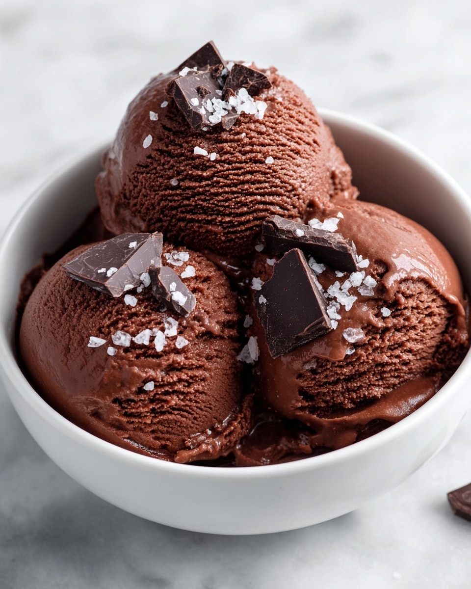 The image shows two close-up views of creamy chocolate ice cream served in a white speckled bowl. The ice cream has a smooth, rich dark brown color with a soft texture and small ridges from scooping. On top of the ice cream are small flakes of dark chocolate and coarse white sea salt crystals that add contrast and texture. The background is a white marbled surface, giving a clean and fresh look. The photo captures the richness and freshness of the ice cream in natural light. Photo taken with an iphone --ar 4:5 --v 7