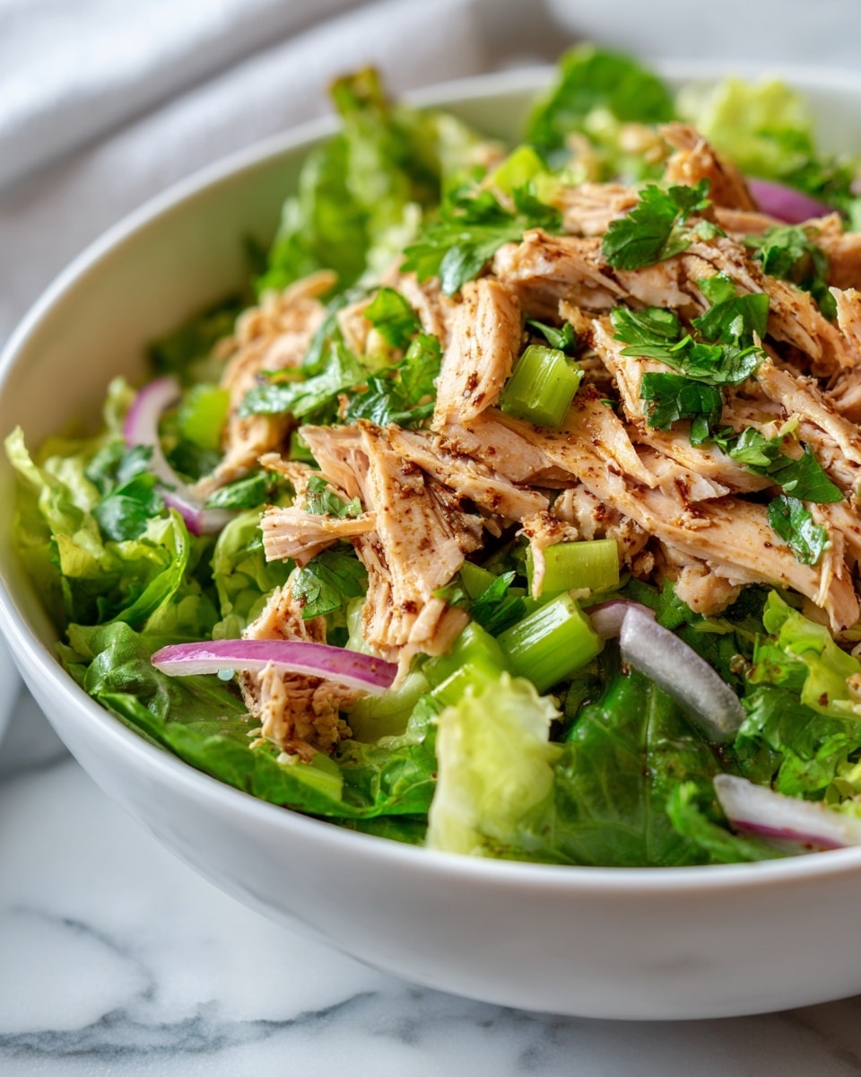 A white bowl filled with a fresh salad showing three main layers: shredded cooked chicken with a lightly grilled, golden-brown look forms the bulk of the dish, mixed with bright green celery pieces cut into small sticks scattered throughout, and topped with thin slices of purple-red onion and fresh, vibrant cilantro leaves adding a leafy texture on top. The textures include the tender, moist chicken, the crisp celery, and the soft leaves of cilantro, all arranged naturally in the bowl sitting on a white marbled surface, photo taken with an iphone --ar 4:5 --v 7