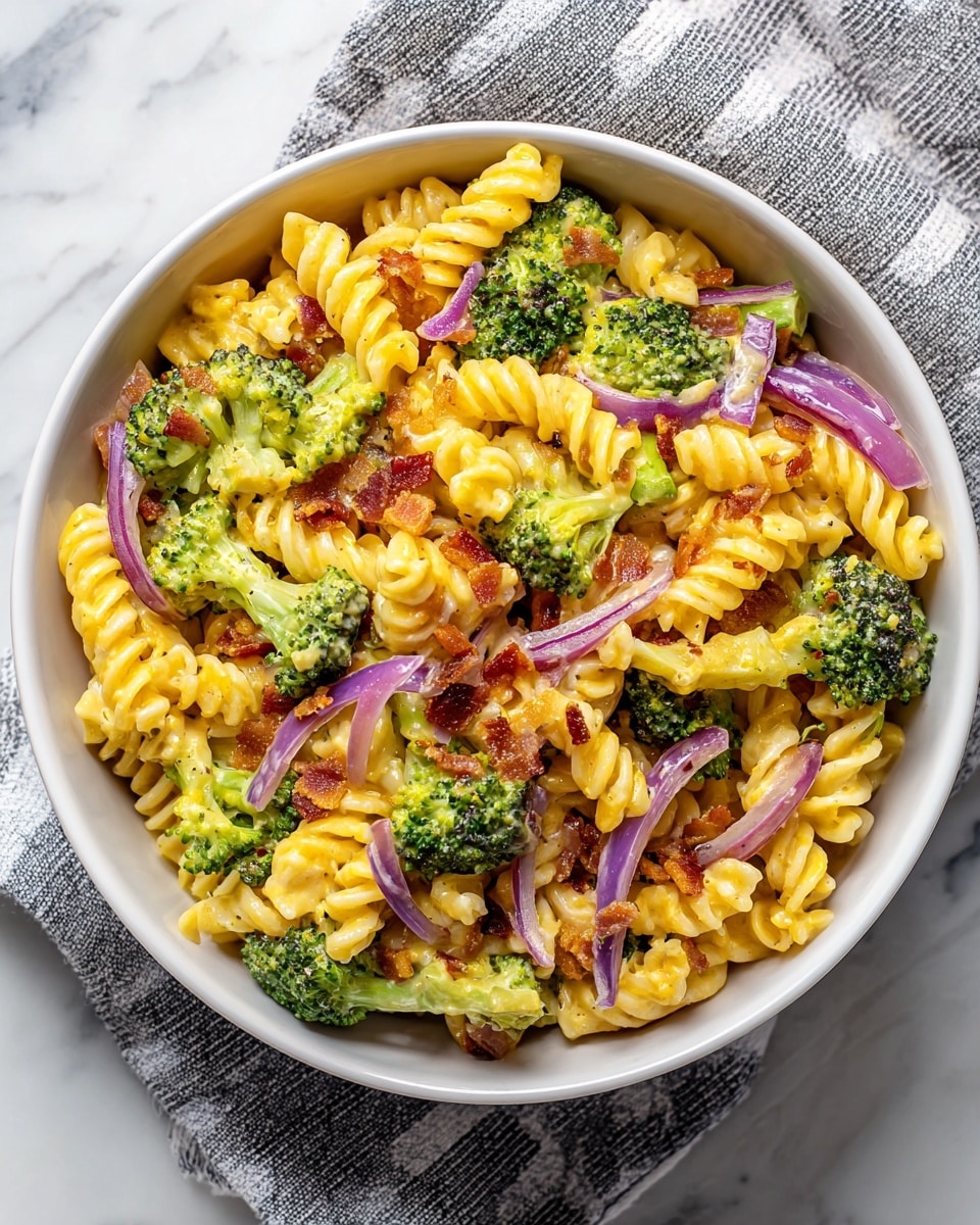 The dish shows a single layer of creamy pasta salad on a white plate, placed on a white marbled texture. The pasta is light yellow and spiral-shaped, mixed evenly with vibrant green broccoli florets that add a fresh, textured look. Scattered thin slices of purple-red onion add color contrast along with small bits of crispy brown bacon sprinkled throughout, creating a mix of smooth, crunchy, and soft textures. The creamy sauce coats the pasta and vegetables lightly, giving a glossy but natural look to the entire dish. Photo taken with an iphone --ar 4:5 --v 7