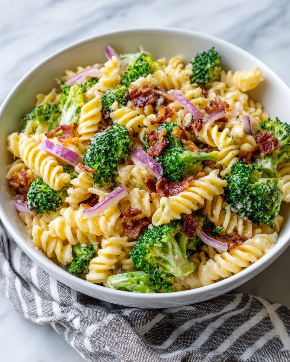 A white bowl filled with a colorful pasta salad featuring three main layers: light yellow spiral macaroni coated in a creamy sauce, bright green broccoli florets with a fresh and slightly rough texture, and thin strips of purple-red onion scattered evenly throughout. Small pieces of crispy brown bacon bits are sprinkled on top, adding texture and contrast. The bowl is placed on a white marbled surface with a gray and white striped cloth partially visible underneath, all creating a fresh, inviting look. Photo taken with an iphone --ar 4:5 --v 7