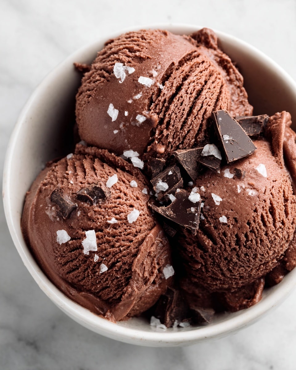 The image shows a white bowl filled with three scoops of creamy, rich dark brown chocolate ice cream. The texture of the ice cream is smooth with some visible ridges from scooping, and it is topped with small chunks of dark chocolate and coarse white sea salt crystals scattered on top. The bowl is placed on a white marbled surface, highlighting the deep color of the ice cream and the contrast of the toppings against the creamy base. photo taken with an iphone --ar 4:5 --v 7