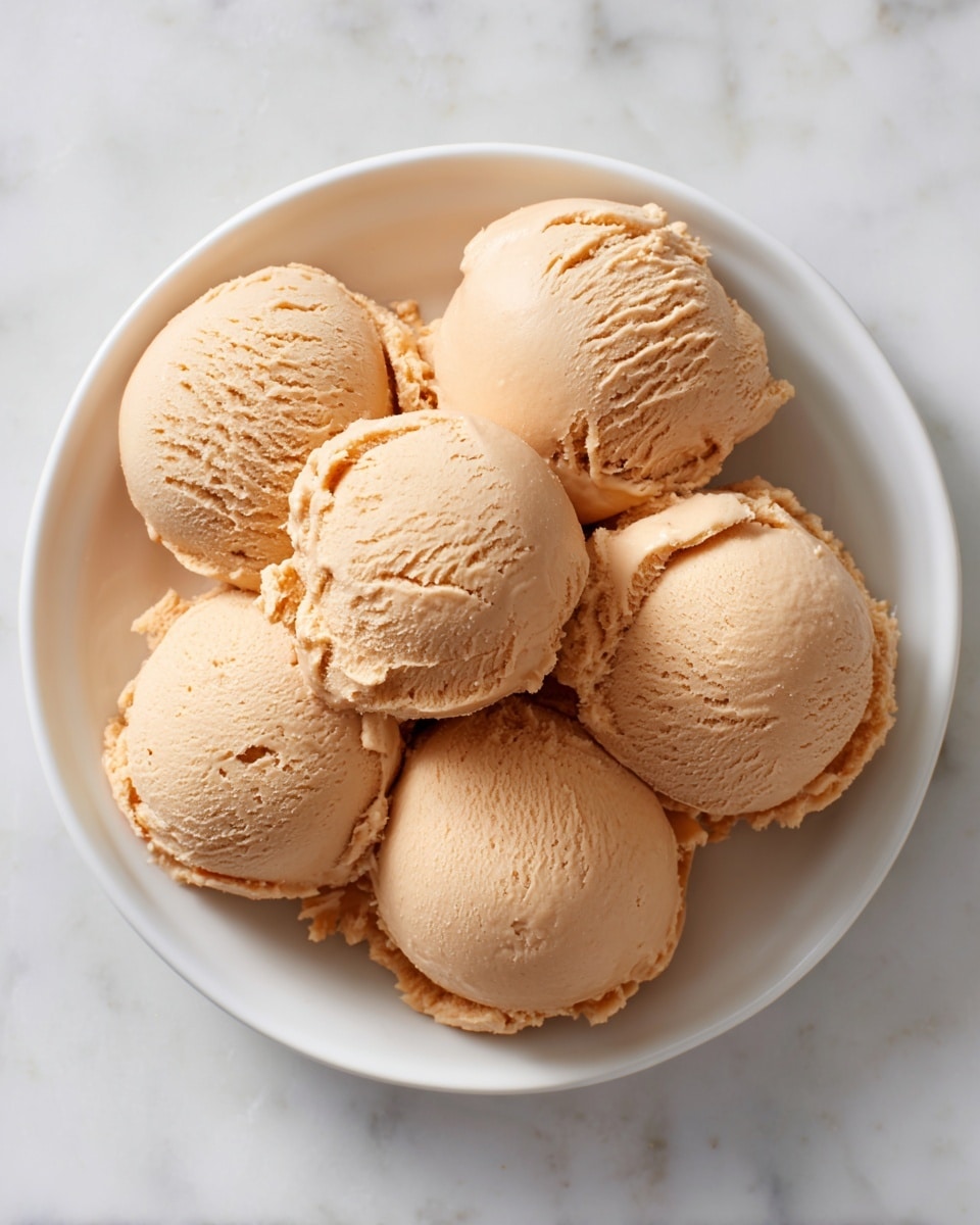 A white round plate sits on a white marbled texture, filled with eight scoops of light brown smooth peanut butter. Each scoop is round with slight ridges and soft texture, stacked closely in the center of the plate, forming a small mound. The soft edges and creamy surface of the scoops give a rich, thick look, with some scoops slightly overlapping. Photo taken with an iphone --ar 4:5 --v 7