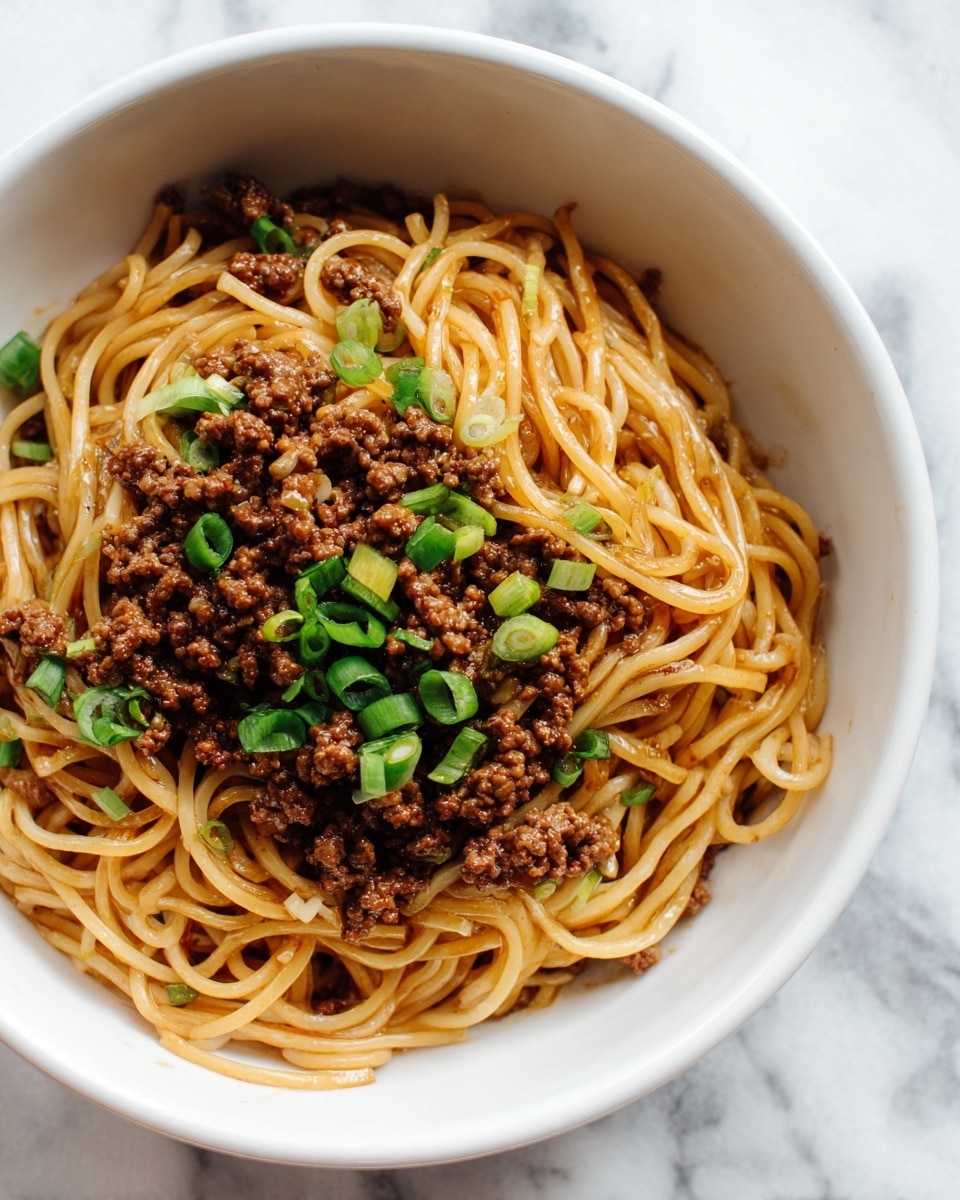 The image shows a close-up of a white plate filled with cooked noodles topped with cooked ground beef. The noodles are light yellow and have a smooth, slightly glossy texture, arranged in loose, thick layers and mixed evenly with the beef. The ground beef is rich brown and crumbly, spread over the top of the noodles in small clusters. Scattered on top are small pieces of green garnish that add a fresh pop of color. The background surface is a white marbled texture. Photo taken with an iphone --ar 4:5 --v 7