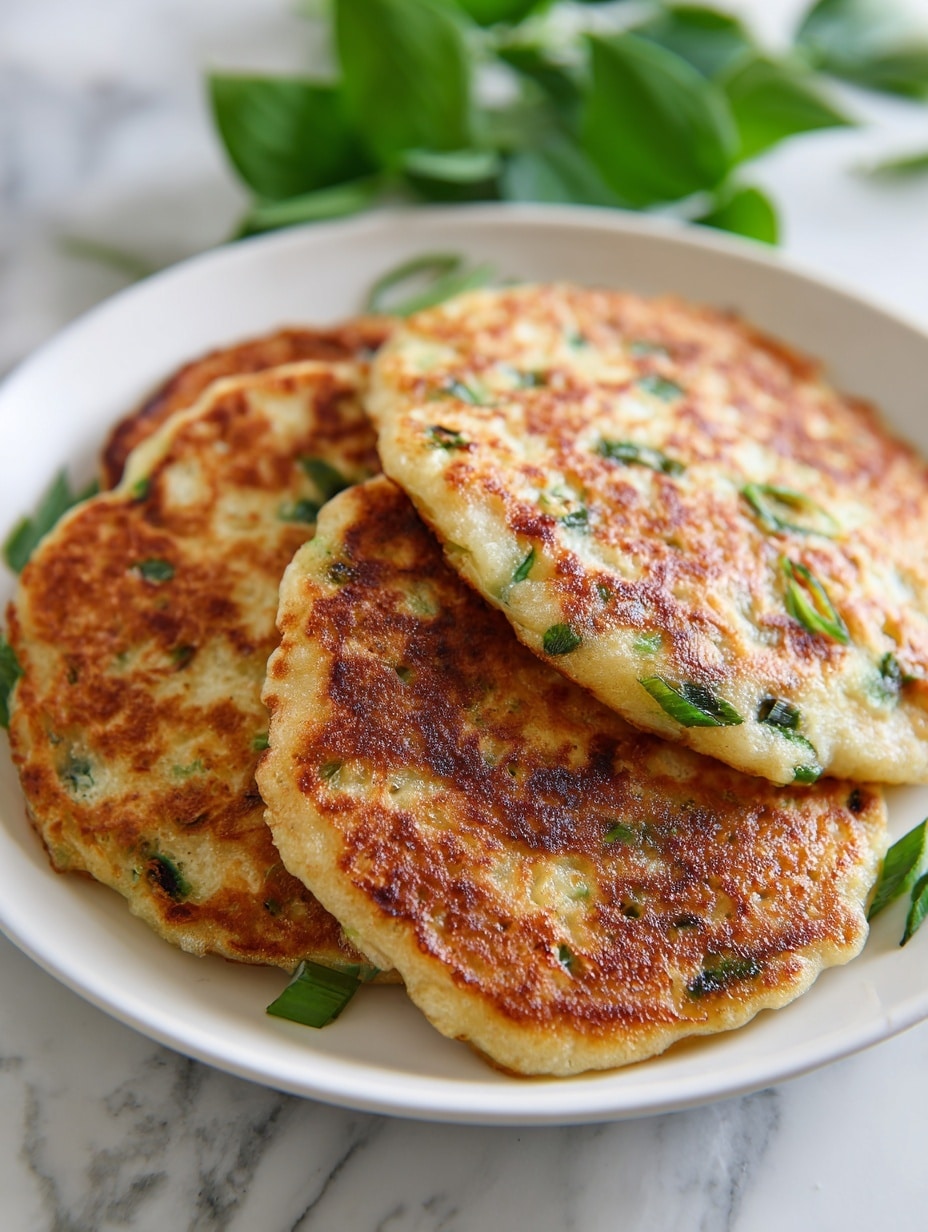A white plate holds three round, thick pancakes stacked slightly overlapping. Each pancake has a golden-brown crispy surface with a slightly bubbly texture and bright green chopped scallions visible inside and on top. The edges are soft and light, contrasting with the toasted browned patches spread unevenly. The plate sits on a white marbled background with long green scallion stalks blurred in the background. photo taken with an iphone --ar 4:5 --v 7